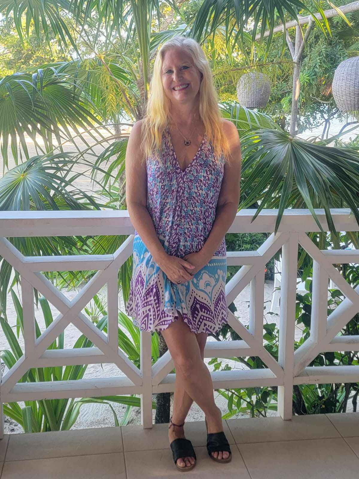 jyll-belize-balcony-purple-dress Jyll Hoyrup standing on a Belize balcony surrounded by tropical plants, smiling in a purple dress